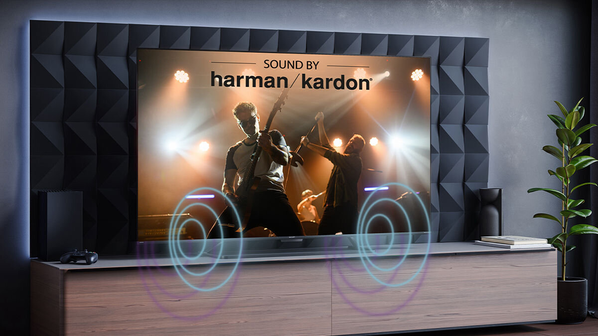 Telewizor wyświetla obraz koncertu z napisem \'SOUND BY harman/kardon\'. W tle widoczna jest nowoczesna szafka RTV i roślina doniczkowa.