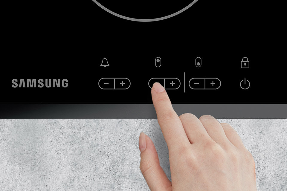 Czarna płyta ceramiczna z dotykowym panelem sterowania, na którym widoczne są symbole dzwonka, plusa i minusa oraz logo SAMSUNG. Dłoń dotyka jednego z przycisków na panelu.