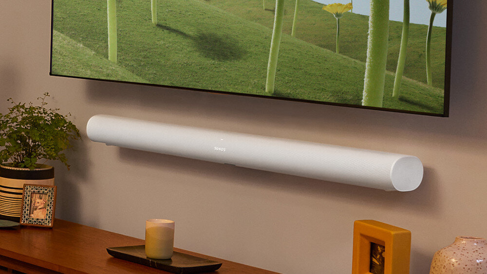 Na ścianie zamontowany jest biały soundbar z widocznym logo \'SONOS\'. W tle znajduje się telewizor z zielonym krajobrazem oraz drewniana półka z rośliną i świecą.