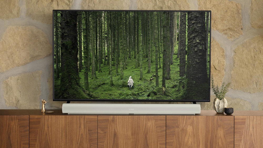 Na drewnianej szafce stoi telewizor wyświetlający obraz lasu z osobą w białym ubraniu. Przed telewizorem znajduje się biały soundbar, a obok niego dekoracje i roślina.