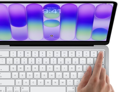 Laptop z widocznym ekranem wyświetlającym kolorowe tło i godzinę 9:41, oraz klawiaturą, na której znajduje się palec naciskający klawisz. Na ekranie widoczna jest data \'Wed Apr 1\'.