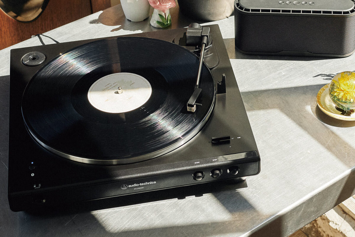 Czarny gramofon z logo audio-technica, na którym odtwarzana jest płyta winylowa. Obok znajduje się głośnik i filiżanka z kwiatem.