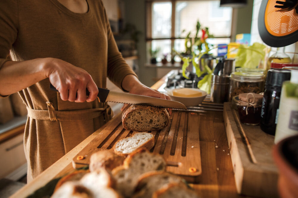 Osoba kroi chleb na drewnianej desce w kuchni, używając dużego noża. W tle widoczne są różne przedmioty kuchenne i rośliny na parapecie.