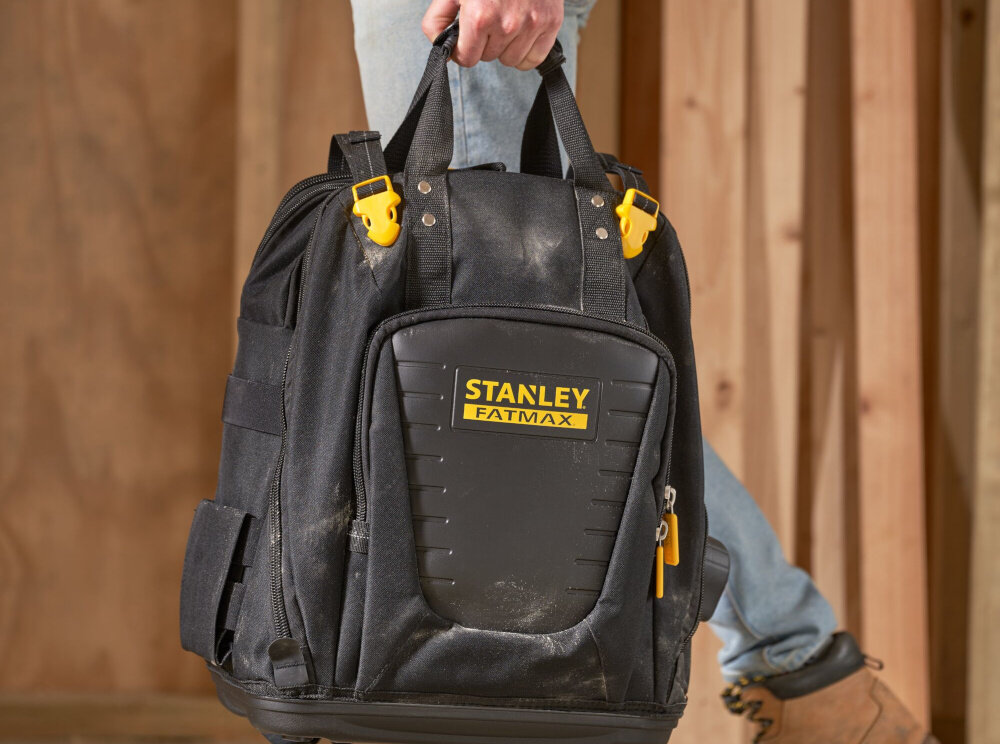 Plecak monterski STANLEY Fatmax Quick Access FMST1-80144 osoba idzie z plecakiem w dłoni trzyma za uchwyt solidny uchwyt górny mocne regulowane szelki noszenie na plecach lub w dłoni elastyczność w transporcie ergonomiczny układ nośny
