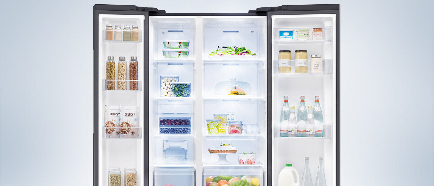 Otwarta lodówka Samsung Side by Side RS62DG5003B1EO z oferty Media Expert, która wyposażona jest w system All-Around Cooling - wizualizacja Otwarta lodówka z widocznymi półkami wypełnionymi różnorodnymi produktami spożywczymi, takimi jak owoce, warzywa, napoje i słoiki. Na drzwiach znajdują się dodatkowe półki z butelkami i pojemnikami.