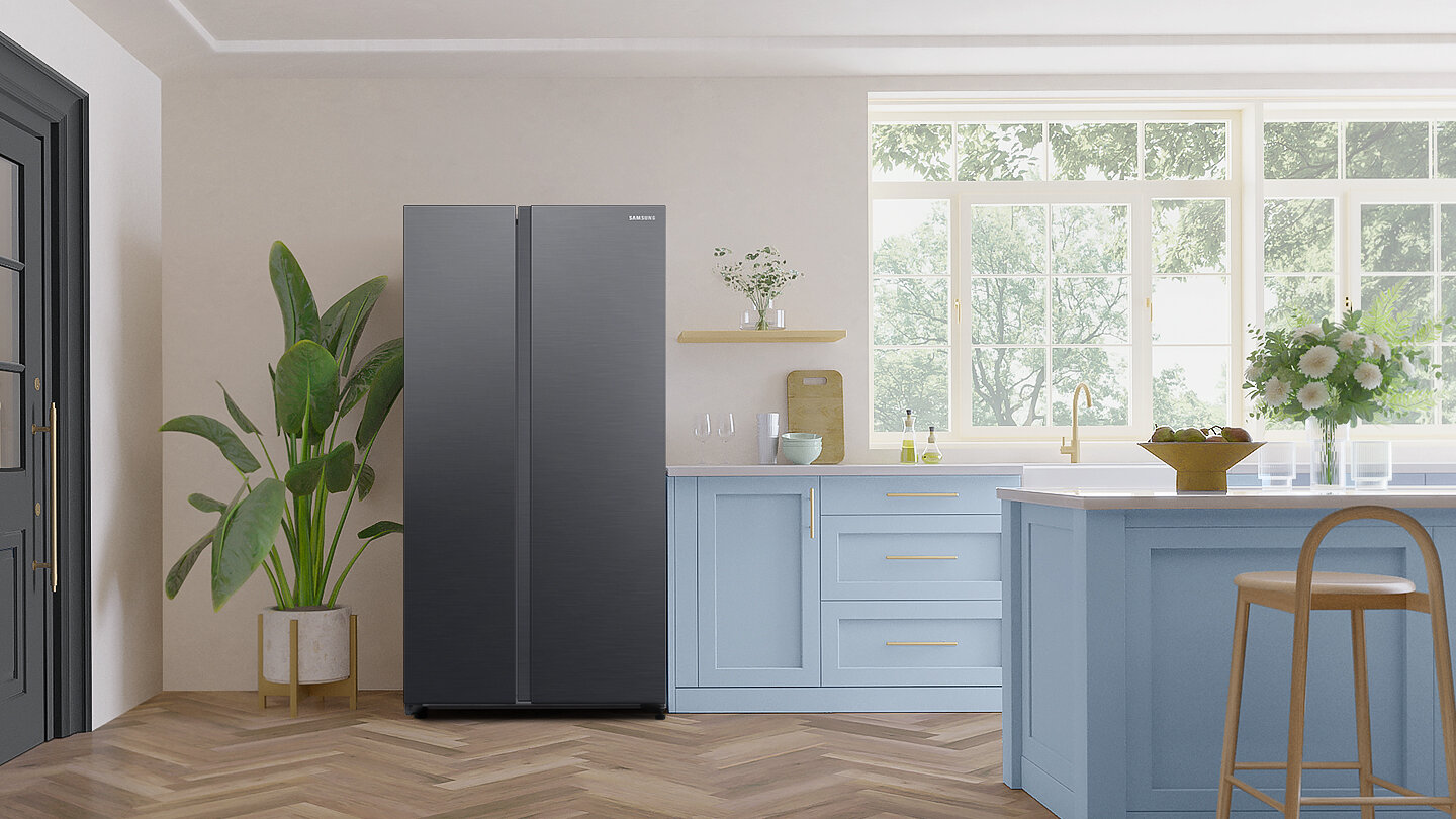 Lodówka Samsung Side by Side RS62DG5003B1EO z oferty Media Expert W kuchni znajduje się lodówka w grafitowym kolorze, obok której stoi roślina w doniczce. Wnętrze jest jasne, z dużym oknem i niebieskimi szafkami.