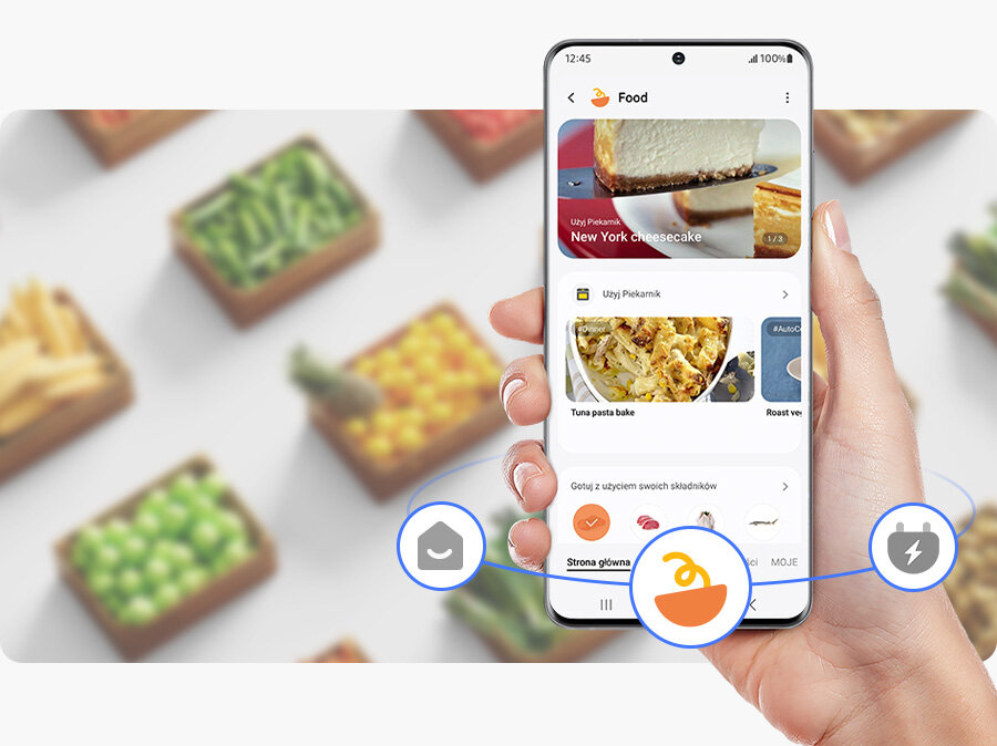 Osoba trzyma smartfon z aplikacją kulinarną, na ekranie widoczny jest przepis na \'New York cheesecake\'. W tle rozmyte pudełka z warzywami.