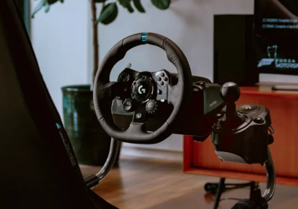 Na obrazie widoczna jest kierownica do gier z logo \'G\' zamontowana na stelażu. W tle znajduje się telewizor z wyświetlonym logo \'FORZA MOTORSPORT\'.