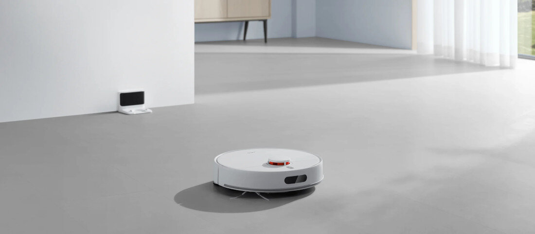 Na jasnej podłodze znajduje się biały robot sprzątający o okrągłym kształcie. W tle widać stację dokującą przy ścianie oraz zasłony przepuszczające światło.