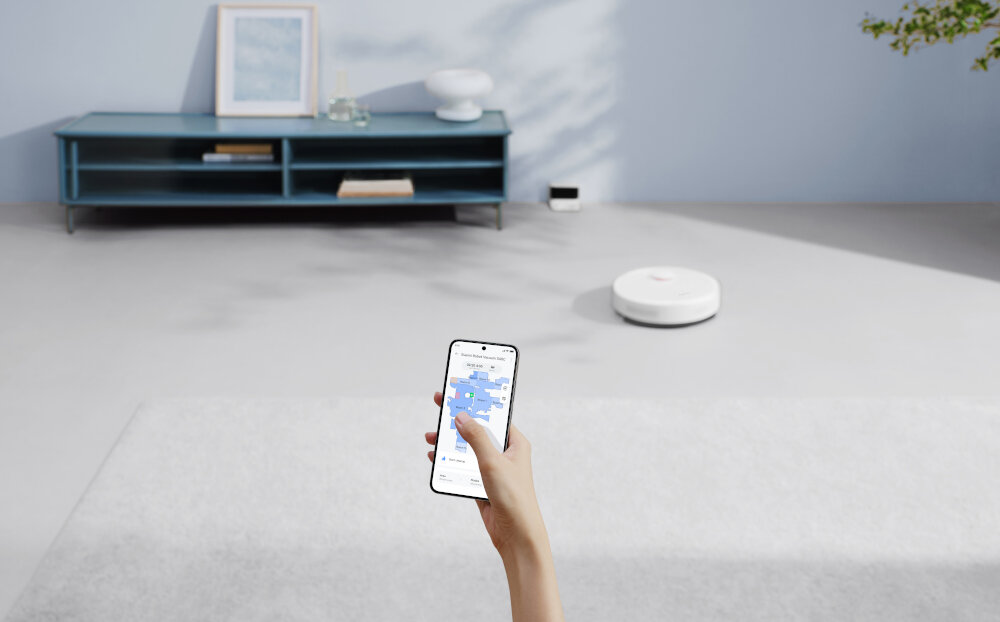 Osoba trzyma smartfon z aplikacją pokazującą mapę pomieszczenia, a w tle widać biały robot sprzątający na podłodze. W pomieszczeniu znajduje się niebieska szafka z dekoracjami.