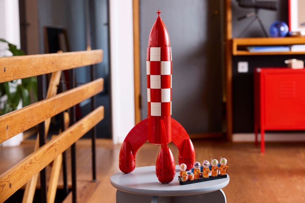 Na obrazie znajduje się model czerwono-białej rakiety z klocków LEGO, stojący na okrągłym stoliku. Obok rakiety ustawione są figurki przedstawiające postacie w skafandrach kosmicznych.