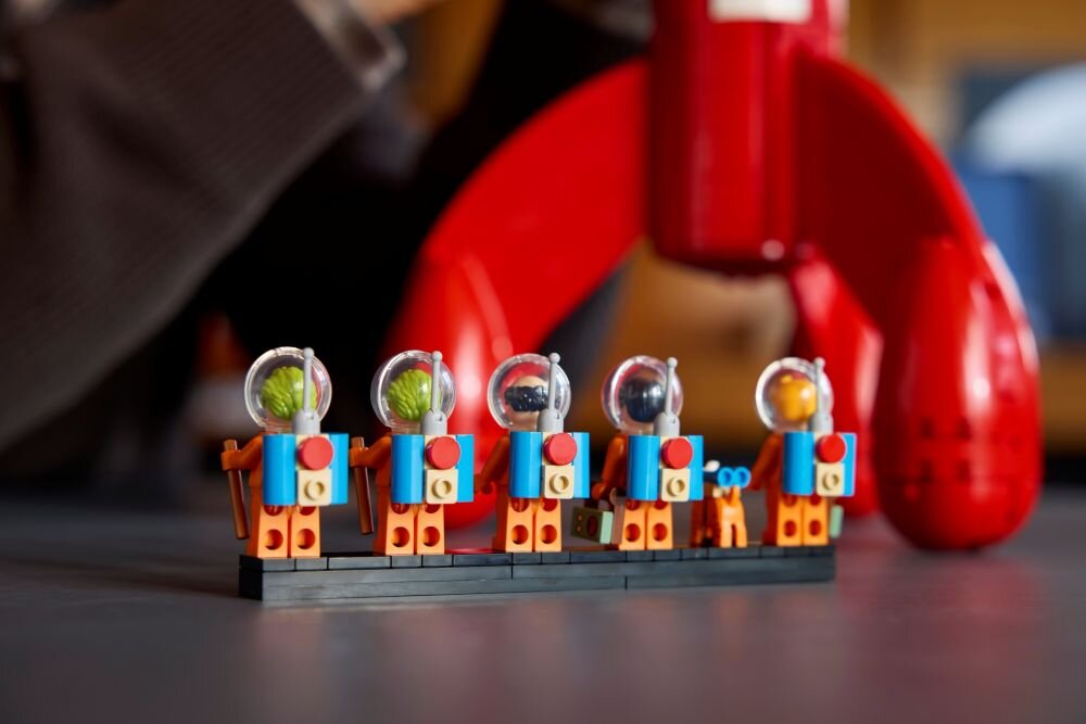 Na obrazie znajduje się zestaw figurek LEGO w skafandrach kosmicznych z przezroczystymi hełmami, ustawionych na czarnej podstawce. W tle widoczna jest duża, czerwona rakieta LEGO.
