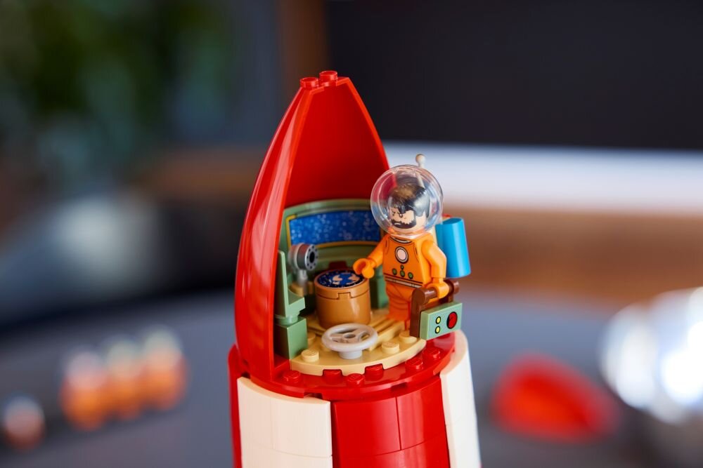 Model rakiety LEGO z otwartą kabiną, w której znajduje się figurka astronauty w pomarańczowym skafandrze. Wnętrze kabiny zawiera detale takie jak panel sterowania i siedzenie.