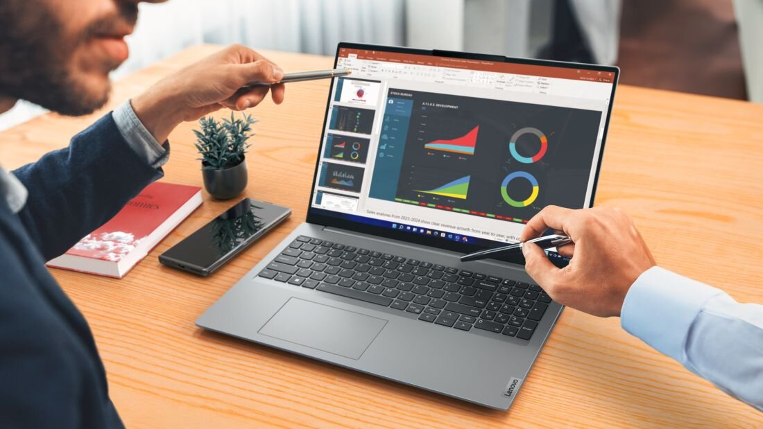 Na drewnianym stole stoi otwarty laptop z wyświetlonym wykresem na ekranie. Dwie osoby wskazują na ekran długopisami, obok laptopa leży smartfon i mała roślina w doniczce.