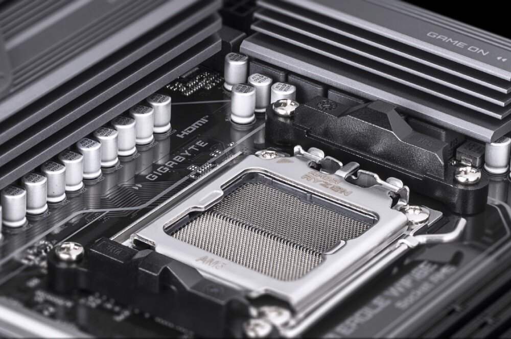Zbliżenie na płytę główną z widocznym gniazdem procesora i napisem \'GIGABYTE\'. W tle widoczne są elementy chłodzenia i kondensatory.