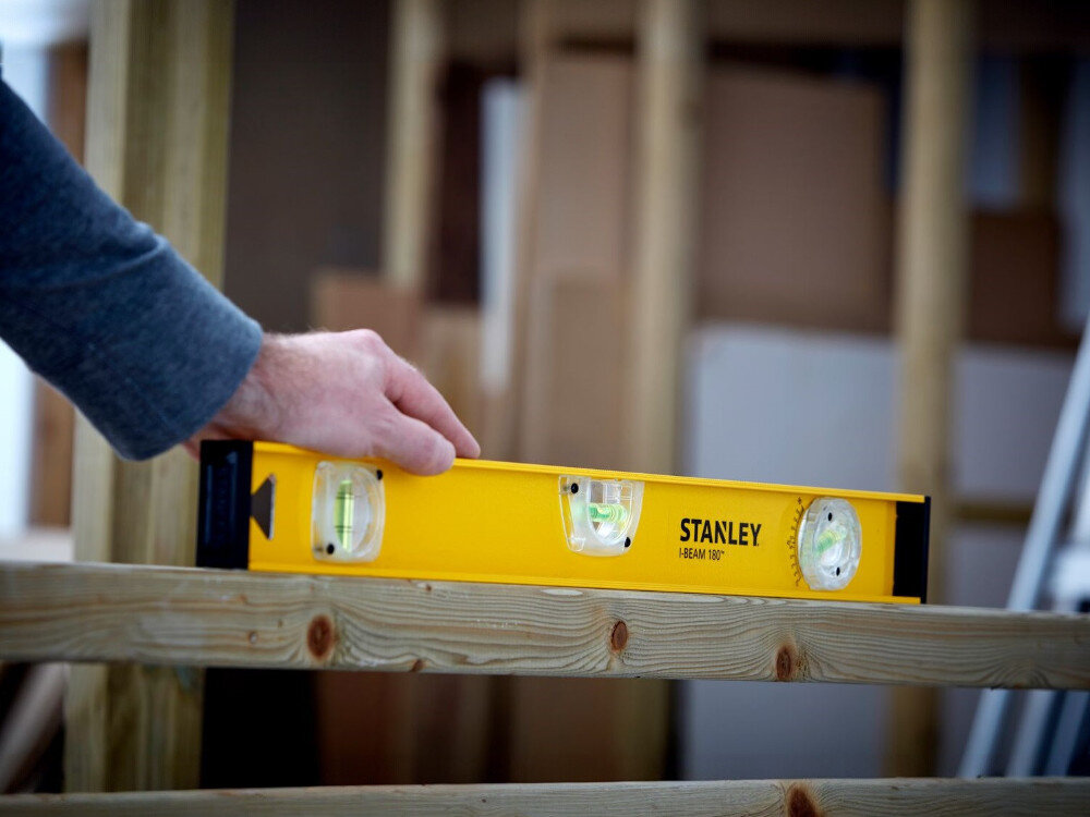 Poziomnica ręczna STANLEY I-Beam 1-42-920, poziom, pion, budowa, pomiar, Dłoń trzymająca żółtą poziomicę Stanley na drewnianej belce podczas prac konstrukcyjnych.