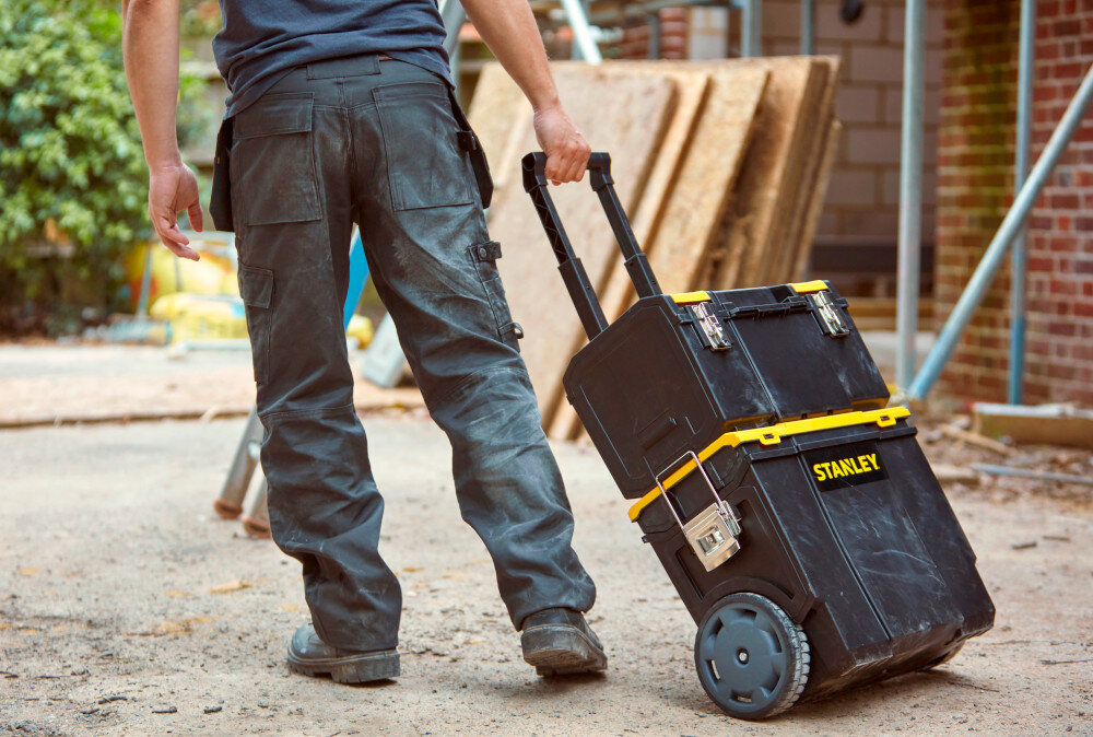 Skrzynka narzędziowa STANLEY 1-70-326 plac budowy osoba ciągnie za sobą skrzynki za pomocą uchwytu w tle drewniane elementy drzewa ceglana ściana wszechstronny system przechowywania z trzech połączonych segmentów wyjmowanej skrzyni z organizerami przestronnej dolnej tacy na większe akcesoria porządku organizacji