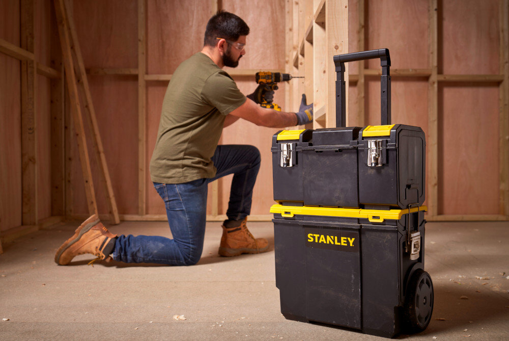 Skrzynka narzędziowa STANLEY 1-70-326 pomieszczenie skrzynki ustawione na podłodze w tle pracuje osoba drewniana ściana dla profesjonalistów majsterkowiczów do użytku w warsztatach na budowach przydomowych garażach wymiary 619 x 475 x 470 mm waga około 4,636 kg