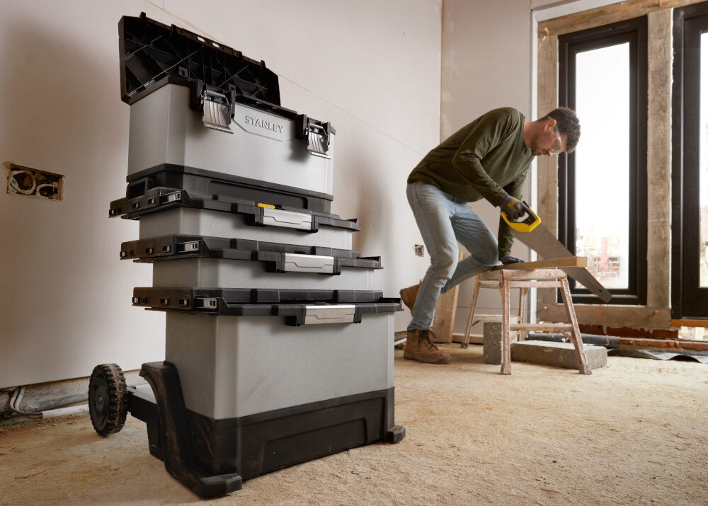 Skrzynka narzędziowa STANLEY FatMax 1-95-622 pomieszczenie drewniana podłoga skrzynki ustawione na podłodze w tle klęczy osoba pracuje drewniana ściana drewniana podłoga okno ruchomy warsztat modułowość mobilność transparent i przechowywanie narzędzi
