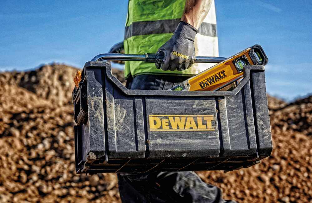 Skrzynka narzędziowa DEWALT DWST1-75654 plac budowy osoba niesie skrzynkę w dłoni skrzynka wypełniona narzędziami metalowy składany uchwyt z ergonomicznym kształtem waga około 2 kg łatwe przenoszenie sprzętu