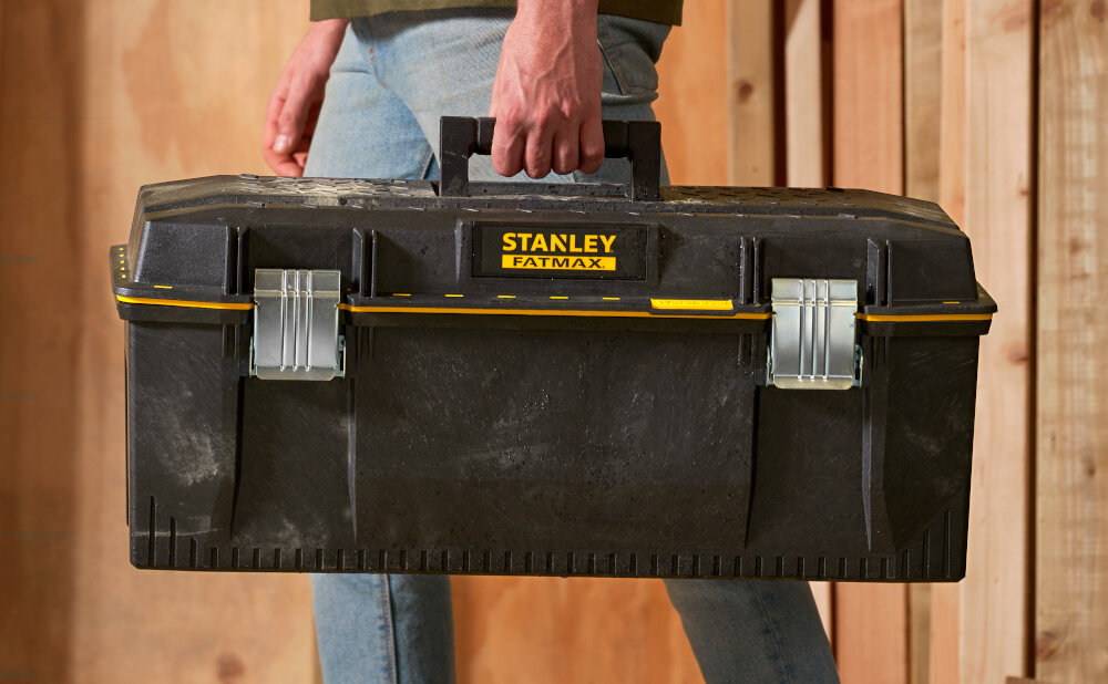 Skrzynka narzędziowa STANLEY 1-92-935 osoba niesie skrzynię w dłoni w tle drewniana ściana duża rękojeść z wysokiej jakości tworzywa sztucznego stabilny chwyt komfort przenoszenia ergonomiczny kształt pewnie leży w dłoni nie ślizga się w czasie transportu
