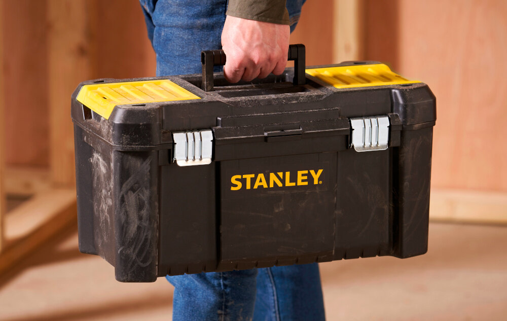 Skrzynka narzędziowa STANLEY Essential STST1-75521 osoba niesie w dłoni skrzynkę w tle drewniana ściana duża rękojeść z wysokiej jakości tworzywa sztucznego stabilny chwyt komfort przenoszenia ergonomiczny kształt pewnie leży w dłoni nie ślizga się w czasie transportu