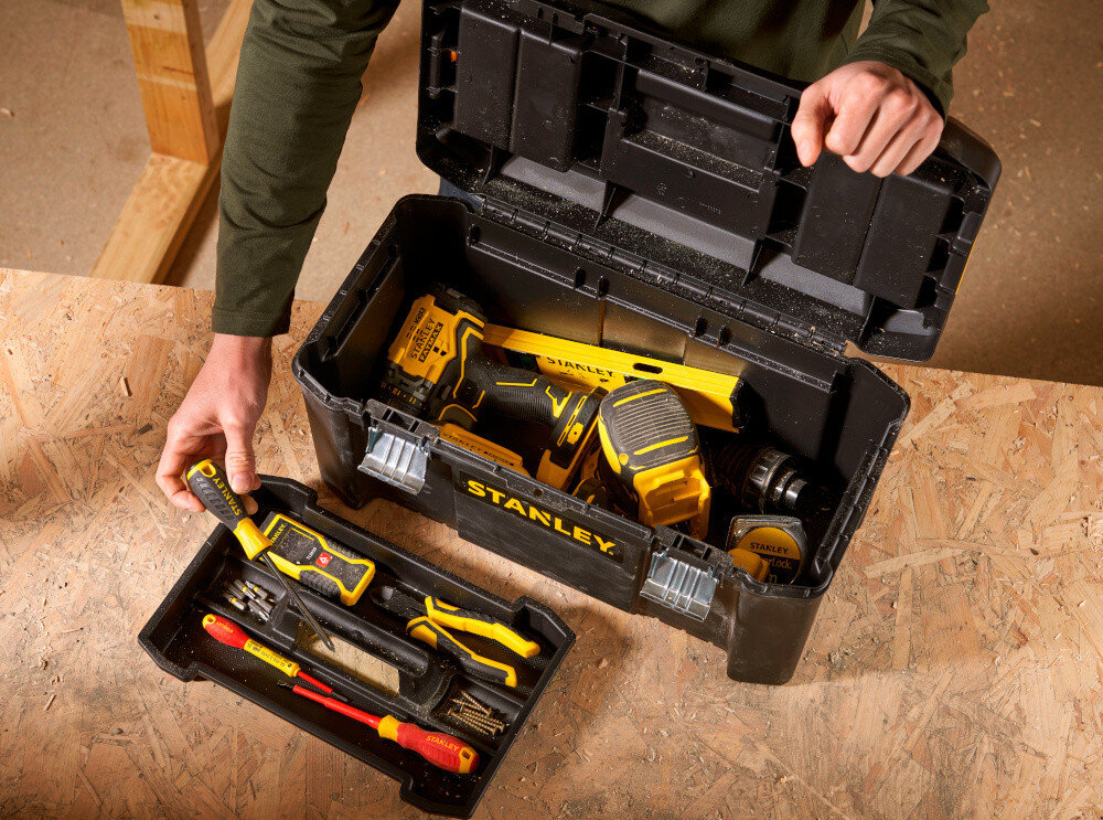 Skrzynka narzędziowa STANLEY Essential STST1-75521 skrzynka na drewnianym blacie obok tacka narzędzia w tle drewniana ściana wygodne przechowywanie narzędzi i akcesoriów porządek w miejscu pracy duża pojemność wnętrza 16,9 litra efektywność komfort