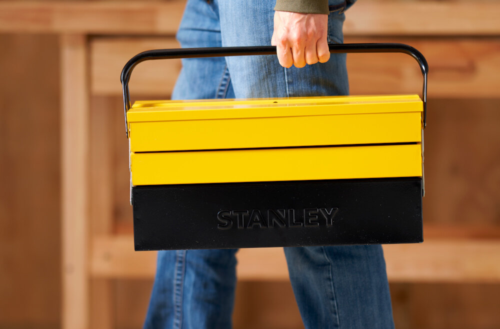 Skrzynka narzędziowa STANLEY S1-94-738 osoba niesie skrzynkę w dłoni w tle drewniana ściana duża dzielona rękojeść udźwig do 35 kg