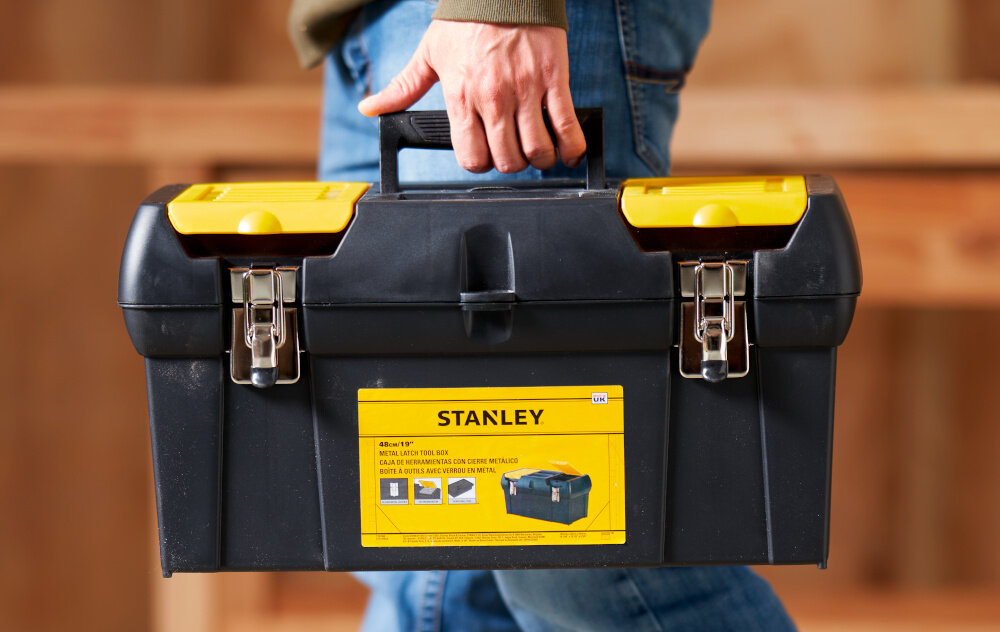 Skrzynka narzędziowa STANLEY 1-92-066 osoba niesie skrzynię w dłoni w tle drewniana ściana duża rękojeść z wysokiej jakości tworzywa sztucznego stabilny chwyt komfort przenoszenia ergonomiczny kształt pewnie leży w dłoni nie ślizga się w czasie transportu