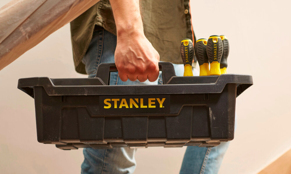 Taca narzędziowa STANLEY STST1-72359 pomieszczenie osoba wchodzi po schodach w dłoni trzyma tacę wypełnioną narzędziami solidny uchwyt do przenoszenia waga 0,83 kg obciążenie do 20 kg organizacja pracy