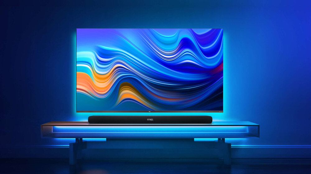 Na obrazie widoczny jest telewizor z kolorowym, abstrakcyjnym wzorem na ekranie, umieszczony na półce z podświetleniem LED. Przed telewizorem znajduje się czarny soundbar z logo TCL.