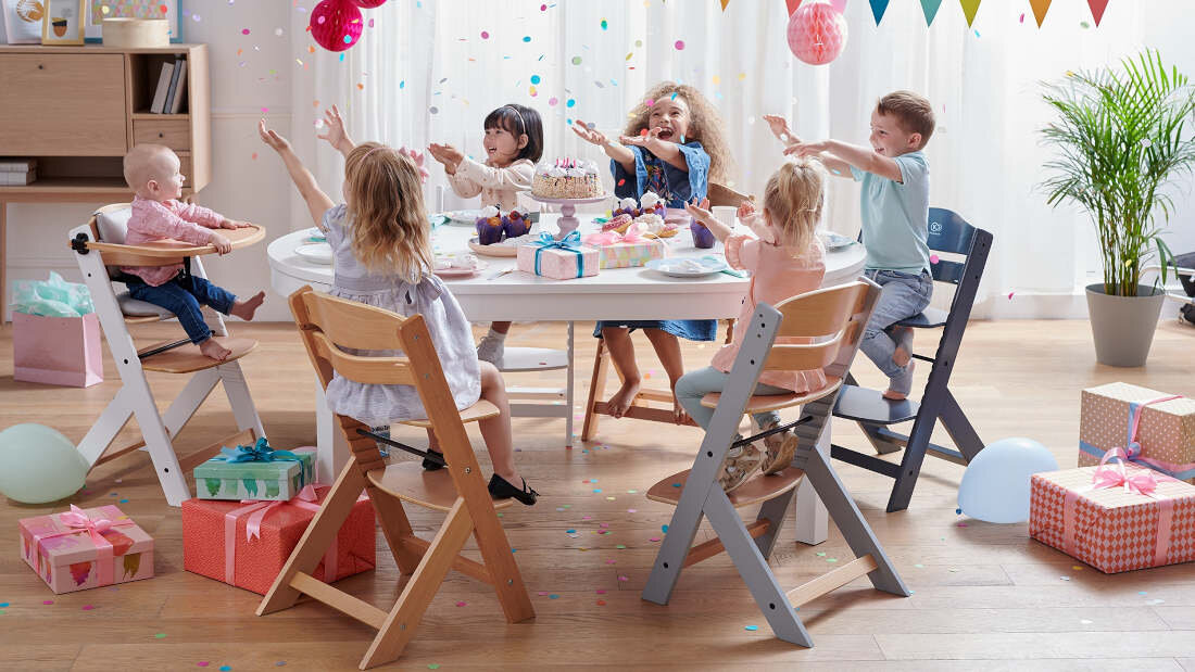 Grupa dzieci siedzi przy stole na krzesełkach do karmienia KINDERKRAFT Enock w kolorze szaro-brązowym, otoczona prezentami i balonami. Na stole znajdują się ciasta i dekoracje, a w tle widać kolorowe girlandy.