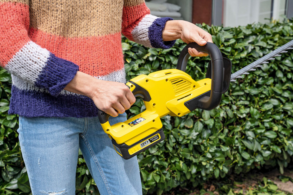 Osoba trzyma żółte nożyce do żywopłotu z widocznym logo KARCHER. W tle znajduje się zielony żywopłot.