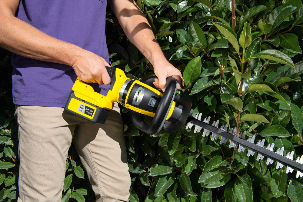 Osoba trzyma żółto-czarne nożyce do żywopłotu z widocznym logo KARCHER, przycinając zielony krzew. Na urządzeniu widoczny jest napis \'Battery Power\'.