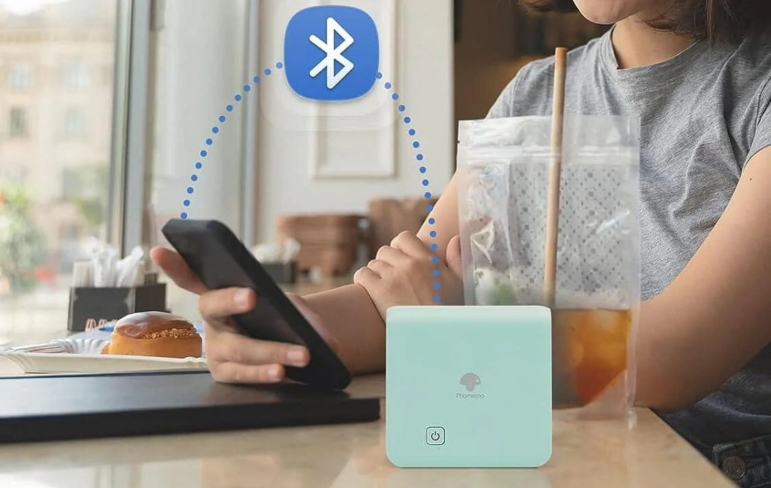 Osoba trzymająca smartfon siedzi przy stole, obok znajduje się mała, biała drukarka z logo \'Phomemo\'. Nad drukarką widoczna jest ikona Bluetooth, sugerująca połączenie bezprzewodowe.