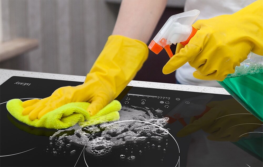 Osoba w żółtych rękawicach sprząta płytę ceramiczną, używając zielonego płynu w sprayu i żółtej ściereczki. Na płycie widoczne są bąbelki mydła, a w rogu znajduje się napis \'ISEASY\'.