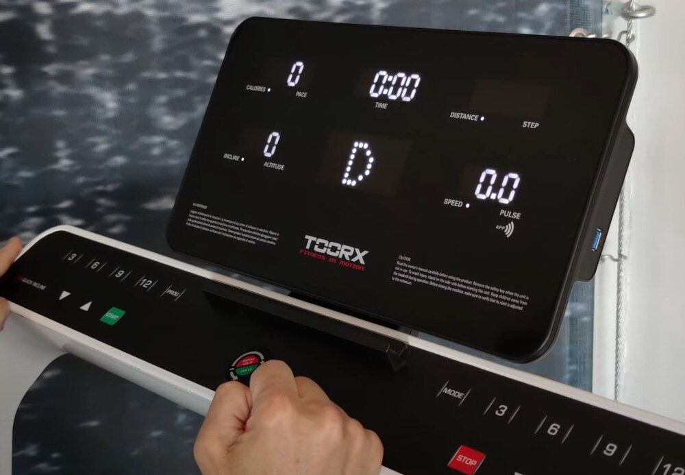 Panel sterowania bieżni elektrycznej z wyświetlaczem pokazującym wartości: kalorie, czas, dystans, prędkość, puls oraz logo TOORX. Widoczna jest ręka obsługująca przyciski na panelu.