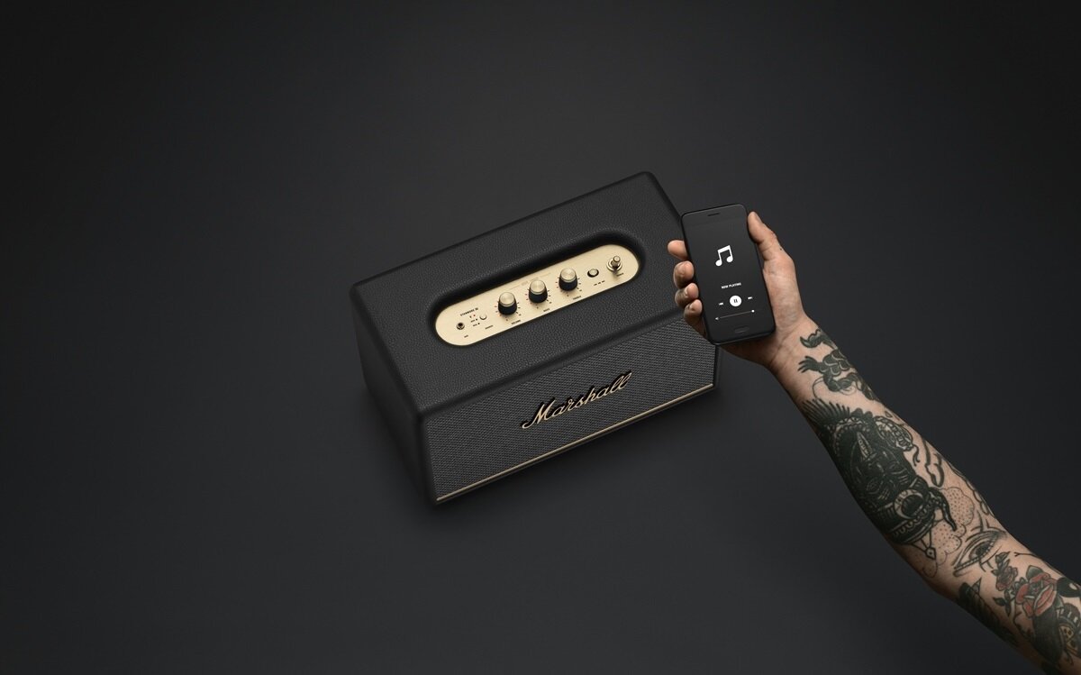 Czarny głośnik z logo \'Marshall\' i złotymi pokrętłami, obok ręka trzymająca smartfon z aplikacją muzyczną na ekranie.