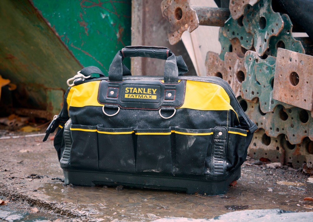 Torba narzędziowa STANLEY 1-93-950 plac budowy torba ustawiona na mokrym podłożu w tle metalowe konstrukcje i elementy dno wykonano z wytrzymałego wodoodpornego tworzywa ochrona przed wilgocią i zabrudzeniami usztywnione panele stabilny kształt