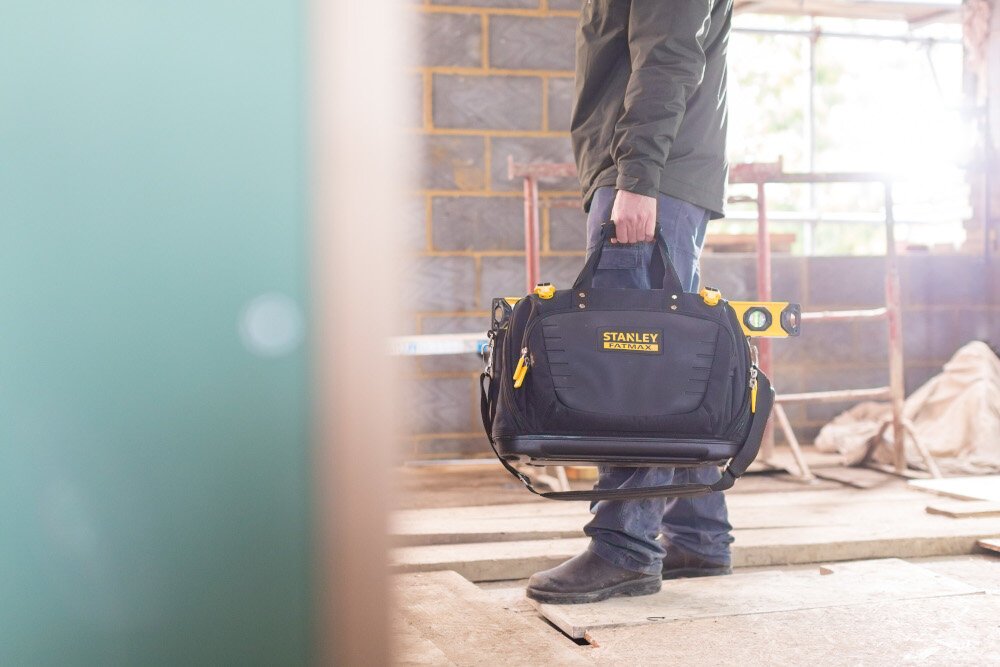 Torba narzędziowa STANLEY FMST1-80147 pomieszczenie osoba niesie torbę w dłoni w tle betonowa ściana rusztowanie okno z odpornego na przetarcia materiału nylon 600 DENIER wzmocniona konstrukcja solidne szwy udźwig do 25 kg