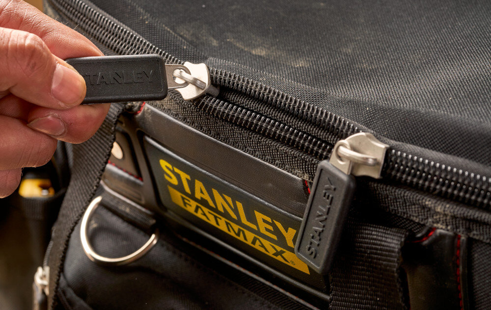 Torba narzędziowa STANLEY FMST1-70749 zbliżenia na zamek osoba trzyma zamek w palcach wykonana z nylonu 600x600 Denier wysoka odporność na przetarcia i rozdarcia wzmocnione szwy dodatkowe nity zamykana jest na zamek błyskawiczny
