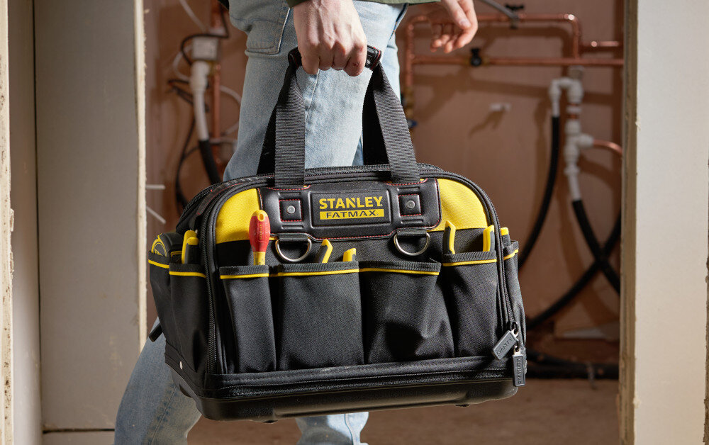 Torba narzędziowa STANLEY Fat Max FMST1-73607 pomieszczenie osoba niesie torbę w dłoni w tle ściana z instalacją wodną wykonana z nylonu 600x600 Denier wysoka odporność na przetarcia i rozdarcia wzmocnione szwy dodatkowe nity
