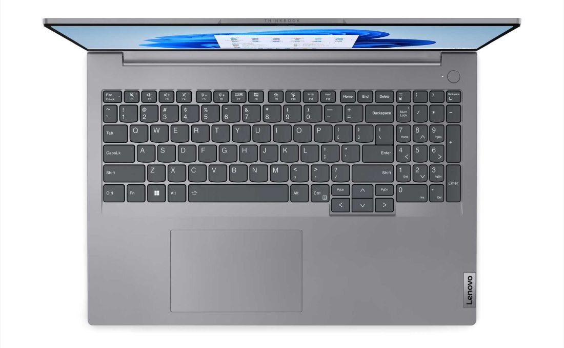 Na obrazie widoczny jest otwarty laptop w kolorze srebrnym z klawiaturą QWERTY i dużym touchpadem. Na ekranie wyświetlany jest interfejs systemu operacyjnego Windows, a na obudowie znajduje się logo \'Lenovo\'.