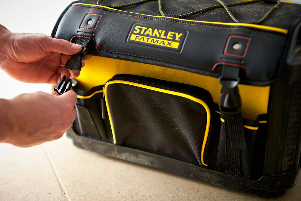 Torba narzędziowa STANLEY Fatmax 1-79-213 pomieszczenie podłoga zamknięta torba osoba trzyma zatrzask w dłoni liczne kieszenie uporządkowane rozmieszczenie narzędzi i akcesoriów szybki dostęp do sprzętu zamykana jest na dwa solidne zatrzaski zabezpiecza zawartość