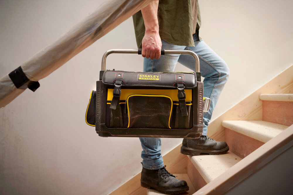 Torba narzędziowa STANLEY Fatmax 1-79-213 pomieszczenie drewniane schody osoba wchodzi po schodach ze skrzynką w dłoni długi metalowy uchwyt pokryty miękką powłoką w miejscu chwytu łatwe przenoszenie torby między stanowiskami pracy