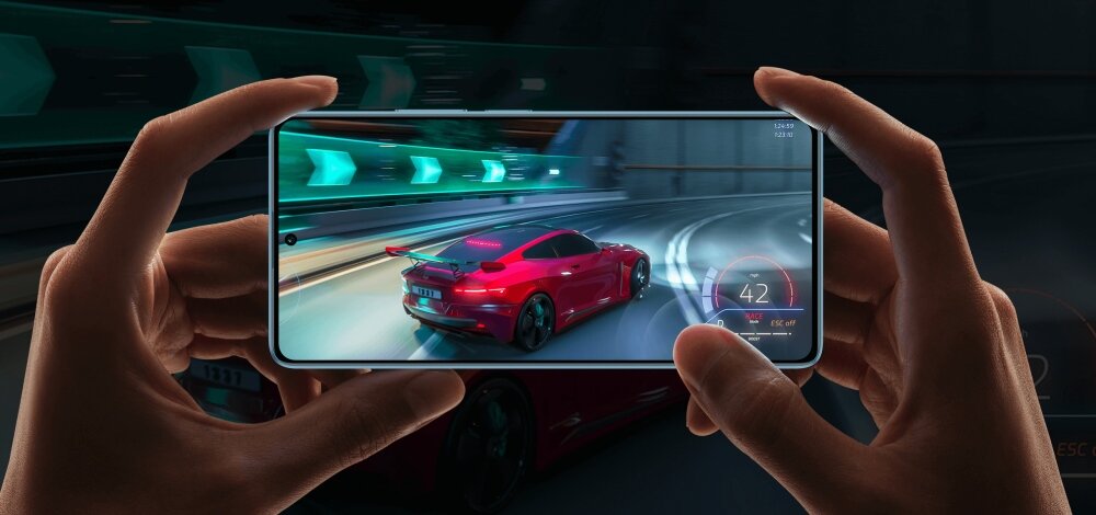Osoba trzyma smartfon, na którym wyświetlana jest gra wyścigowa z czerwonym samochodem na torze. W tle widać rozmyty obraz tunelu z zielonymi światłami.