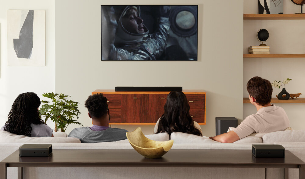 Cztery osoby siedzą na kanapie w salonie, oglądając film na telewizorze zamontowanym na ścianie. Na szafce pod telewizorem znajduje się soundbar, a w pomieszczeniu widoczne są elementy dekoracyjne i rośliny.