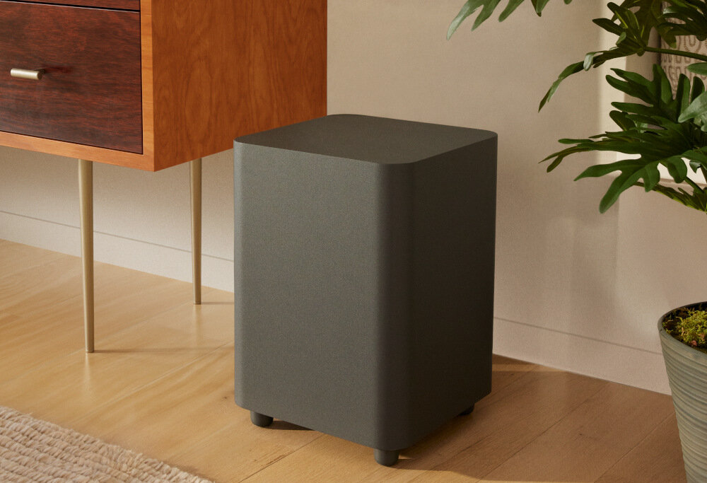 Na drewnianej podłodze stoi czarny subwoofer o zaokrąglonych krawędziach. Obok znajduje się drewniana szafka i roślina w doniczce.