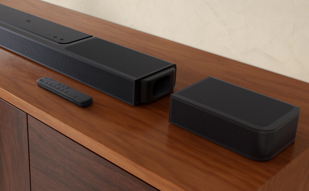 Na drewnianej powierzchni znajduje się czarny soundbar, obok którego leży pilot oraz dodatkowy głośnik. Urządzenia mają nowoczesny design i są wykonane z ciemnego materiału.