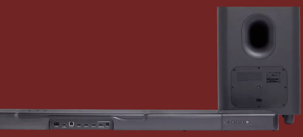 Na obrazie widoczny jest czarny soundbar z kilkoma portami z tyłu oraz bezprzewodowy subwoofer z otworem bass-reflex. Tło jest jednolite, ciemnoczerwone.